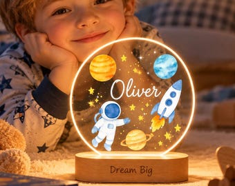 May include: A circular acrylic night light featuring a space theme with a watercolor astronaut, rocket ship, planets, and stars. The name Oliver is printed in white script. The light sits on a wooden base engraved with the text Dream Big.