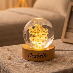 May include: A clear glass sphere containing yellow flowers, resting on a wooden base. The base is inscribed with the name "Isabelle" in a cursive font. The sphere is illuminated from within, creating a warm glow.