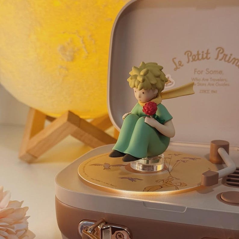 K&ouml;nnte beinhalten: Eine Figur des kleinen Prinzen, der eine rote Rose h&auml;lt und auf einem Plattenspieler sitzt. Der Plattenspieler ist beige und wei&szlig; mit einer goldfarbenen Schallplatte. Der Hintergrund umfasst eine gelbe mondf&ouml;rmige Lampe und den Text "Le Petit Prince".