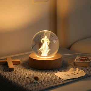 May include: A clear glass sphere with an illuminated figure inside, set on a wooden base. The sphere is lit from within, creating a warm glow. A wooden cross, a small book, a daisy, and a note that says "Thank you God, for that prayer" are also present.