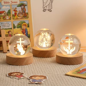 Puede incluir: Tres esferas de vidrio iluminadas con imágenes religiosas, cada una sobre una base de madera. Las esferas presentan una cruz, manos rezando y una representación de la Sagrada Familia. El brillo cálido emana desde el interior.