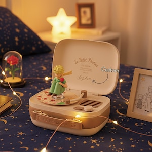 May include: A beige and brown music box with a figurine of The Little Prince holding a red rose. The box has a turntable design with the text "Le Petit Prince" and "Custom" on the lid. A star-shaped lamp and fairy lights are in the background.