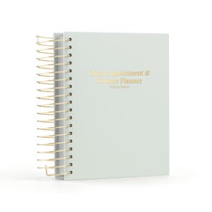 Peut inclure: Un agenda à reliure spirale vert sauge clair avec une spirale dorée. La couverture présente un texte doré imprimé indiquant Client Appointment & Finance Planner et Wellness Planners. L'agenda possède une couverture unie et épurée.