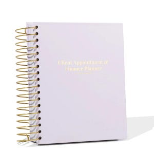 Puede incluir: Una agenda lila con espiral y encuadernación metálica dorada. La portada presenta el texto "Client Appointment & Finance Planner" en letras doradas. La agenda está diseñada para organizar citas y finanzas.