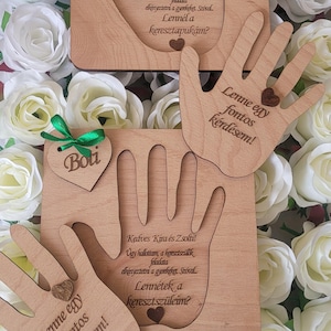 Puede incluir: Placas de madera de color marrón claro con forma de manos, con texto grabado, detalles de corazón y etiquetas en forma de corazón con nombres. Las placas están sobre un lecho de rosas blancas. El texto está en un idioma extranjero.