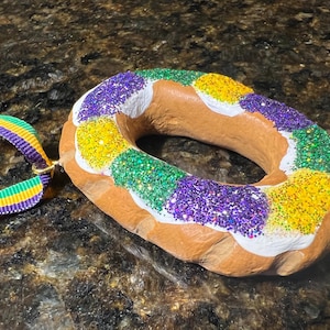 May include: A brown, glazed donut with purple, green, and yellow sprinkles. The donut is decorated with white icing and has a hole in the center. The donut is hanging from a purple, green, and gold ribbon.