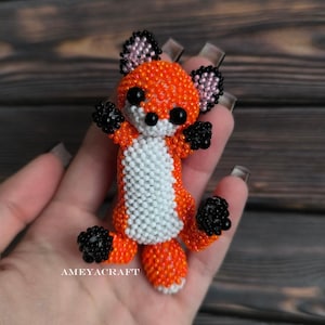 Könnte beinhalten: Eine handgefertigte Fuchsskulptur aus Perlen mit orangefarbenen, schwarzen, weißen und rosa Akzenten. Der Fuchs hat schwarze Perlenaugen, schwarze Pfoten und eine weiße Brust. Die Ohren sind rosa und schwarz. Die Figur trägt den Text "AMEYACRAFT".