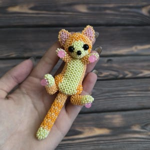 May include: A handmade beaded cat figurine in orange, yellow, and pink. The cat has black eyes and pink accents on its paws and ears. The figurine is held in a hand against a wooden background.