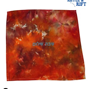 May include: A square, tie-dye textile in shades of red, orange, and brown. The fabric features the Hebrew text "Shabbat Shalom" in light blue. The upper right corner has the text "Ripple & Rift Studio" in blue.
