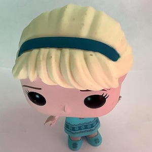May include: A Funko Pop figure of a character with a light blonde hairstyle, a teal headband, and a teal dress. The figure has large black eyes and is wearing teal shoes. The figure is a collectible item.