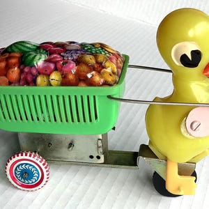 Peut inclure: Un canard en plastique jaune tirant un chariot vert rempli de fruits colorés. Le canard a une aile rose et des yeux noirs. Le chariot a un cadre en métal et des roues.