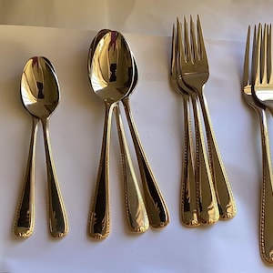 May include: A selection of stainless steel cutlery with gold-coloured detailing. The set includes a knife, spoons, and forks. The cutlery features a decorative pattern on the handles. The items are arranged on a white surface.