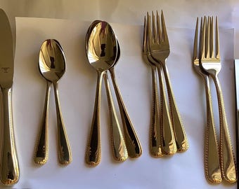 May include: A collection of stainless steel silverware with gold-colored accents. The set includes a knife, spoons, and forks. The silverware has a decorative pattern on the handles. The items are arranged on a white surface.