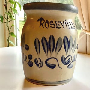 May include: A beige Roseville pottery jar with blue floral designs and the word "Roseville" in black script. The jar has a dark blue rim and base, and the floral pattern includes stylized leaves and swirls.