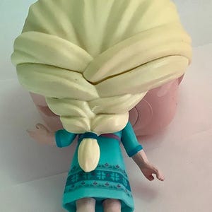 May include: A figurine of a character with blonde hair styled in a braid, wearing a teal dress with a floral pattern. The figure has a light pink face and teal shoes. The image is taken from the back.