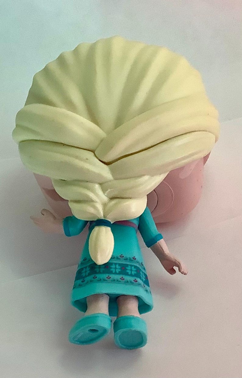 May include: A figurine of a character with blonde hair styled in a braid, wearing a teal dress with a floral pattern. The figure has a light pink face and teal shoes. The image is taken from the back.