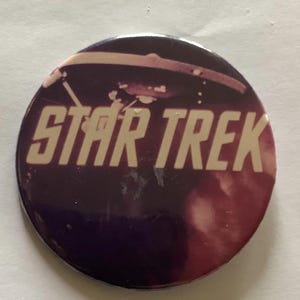 May include: A round button with the words "STAR TREK" in white block letters. The background is a gradient of purple and red, with a blurred image of a spaceship. A collectible item.