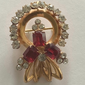May include: A vintage gold-tone brooch with a circular design. The brooch features a ring of clear rhinestones, with three large, rectangular red stones and smaller clear stones. The brooch has a pin closure on the back.
