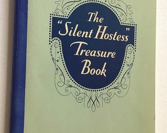 May include: A hardcover book titled The Silent Hostess Treasure Book with a light green cover, a dark blue spine, and a central dark blue shield-shaped emblem featuring decorative black scrollwork and the GE logo in a small circle at the top.
