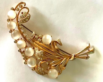 May include: A gold-toned brooch in a curved, floral design. The brooch features a series of large, round, off-white faux pearls and small, clear rhinestones. The brooch has a pin closure on the back.