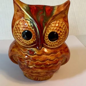 Puede incluir: Figura de búho de cerámica con acabado brillante. El búho presenta ojos grandes y texturizados y un patrón de plumas detallado en tonos marrones, naranjas y rojos. Los ojos del búho son negros y la figura tiene un pequeño acento rojo en el pecho.