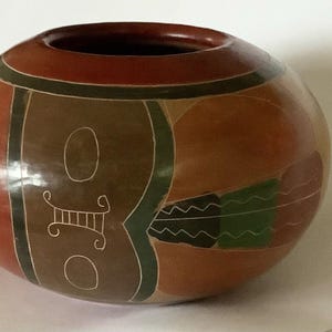 May include: A handcrafted, round ceramic vase with a wide opening. The vase features a reddish-brown base colour with brown, green, and cream-coloured geometric designs. The vase is approximately 20 cm in diameter.