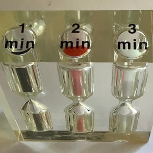 May include: A clear acrylic timer with three hourglasses, each marked with a number and "min" for minutes. The first hourglass is black, the second has red sand, and the third is empty. The timer is designed to measure 1, 2, and 3 minutes.