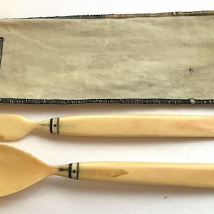 Puede incluir: Un juego de cubiertos de viaje de color marfil con un tenedor y una cuchara, ambos con ribetes negros. Los cubiertos están en una bolsa de tela beige con ribetes negros y una etiqueta que dice "The Sterling Silver Company, New York".