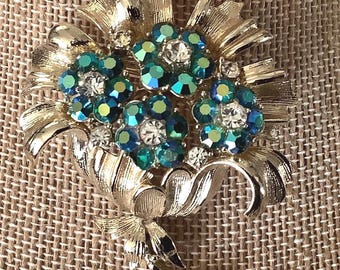 May include: A gold tone brooch with a cluster of green and clear rhinestones in the shape of flowers. The brooch has a leaf design.