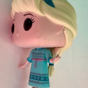 May include: A Funko Pop figure of Elsa from Frozen. The figure has a large head, black eyes, and blonde hair. She wears a teal dress with a pink belt and blue shoes. The figure is approximately 4 inches tall.