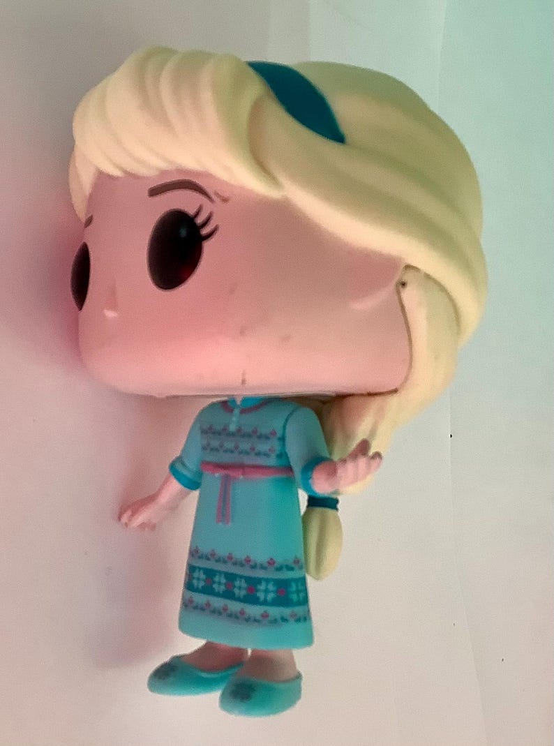 May include: A Funko Pop figure of Elsa from Frozen. The figure has a large head, black eyes, and blonde hair. She wears a teal dress with a pink belt and blue shoes. The figure is approximately 4 inches tall.