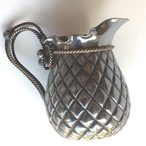 May include: A silver creamer with a pineapple-shaped body and a rope-like handle with brass accents.