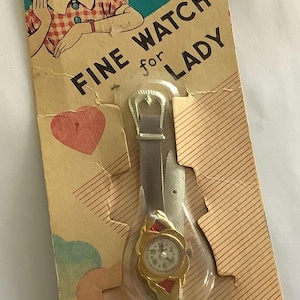 Puede incluir: Reloj vintage "Fine Watch for Lady" en su embalaje original. El reloj tiene una caja dorada, una esfera blanca y detalles rojos. El embalaje presenta una ilustración de dibujos animados de una niña y el texto "Made in Japan".