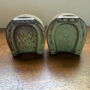 May include: A pair of ceramic salt and pepper shakers shaped like horseshoes. The shakers feature a mottled light green glaze with brown accents along the edges and a series of small holes at the top for dispensing seasoning.