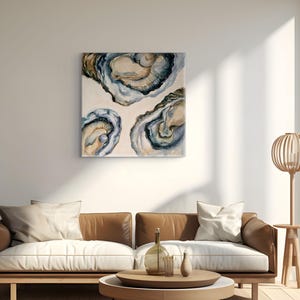 以下が含まれることがあります： 青、茶、クリーム色のトーンで描かれた3つの開いた牡蠣の絵画が特徴の正方形のウォールアート。1つの牡蠣には小さな丸い白い真珠が含まれています。このアート作品は、茶色の革製ソファの上の白い壁に飾られています。