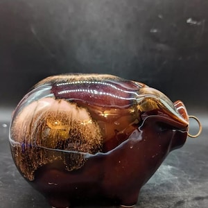 Pode incluir: Um cofrinho de cerâmica marrom com um padrão de espiral de esmalte marrom, dourado e branco. O cofrinho tem um pequeno anel de metal na lateral para inserir moedas.
