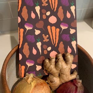 May include: A brown notebook with a colorful pattern of vegetables and roots. The notebook is sitting on a brown bowl with a yellow onion, a red onion, a ginger root, and a few cloves of garlic.