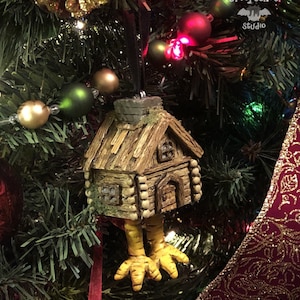 May include: A whimsical Christmas tree ornament shaped like a rustic cabin with chicken legs. The cabin is brown with a chimney and small windows. The chicken legs are yellow. The ornament hangs on a Christmas tree with green needles and colorful ornaments.