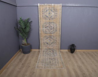 May include: A long, rectangular, vintage-style rug with a beige background and repeating blue geometric patterns. The rug is displayed vertically against a gray wall, with a potted plant and a decorative vase on either side.