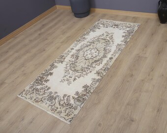 May include: A rectangular, cream-colored rug with an ornate, floral pattern in shades of gray and brown. The rug is placed on a light-colored wooden floor, with a dark gray wall in the background. The rug's design features a central medallion and border.