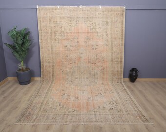 Puede incluir: Una alfombra vintage grande y rectangular con un aspecto descolorido y desgastado. La alfombra presenta un diseño central en tonos melocotón y beige, con detalles de borde más oscuros. La alfombra se exhibe contra una pared gris.