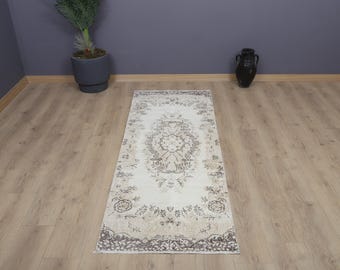 May include: A rectangular, cream-colored rug with an intricate, floral-inspired pattern in shades of beige and gray. The rug is placed on a light-colored wooden floor, with a dark vase and a potted plant in the background.