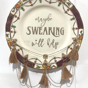 May include: A decorative ceramic plate featuring a cream centre with the text "maybe SWEARING will help" in black lettering. The rim has a dark purple and floral pattern. Gold-toned chains and five light brown tassels hang from the plate edge.