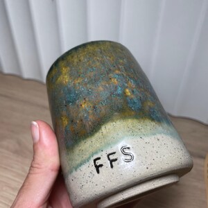 May include: A handmade ceramic tumbler with a speckled, off-white base and a gradient of colours on the upper portion, including teal, gold, and brown. The letters "FFS" are stamped near the base. The tumbler is held by a hand.