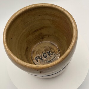 May include: A handmade ceramic mug with a tan exterior and a dark blue stripe near the base. The interior is a lighter tan colour. The word "FUCK" is stamped on the base of the mug.