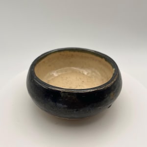 May include: A small, handmade ceramic bowl. The exterior is dark and glossy, while the interior is light beige. The bowl has a rounded shape and a slightly textured surface, ideal for serving dips or small portions.