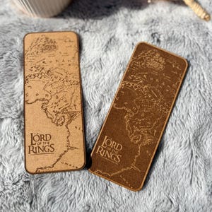 Puede incluir: Dos marcapáginas rectangulares de madera con un diseño de mapa y el texto "LORD OF THE RINGS". Uno es marrón claro, el otro es marrón oscuro. Un jarrón blanco y flores secas en el fondo.