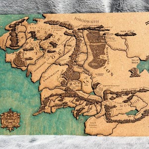 May include: A layered wooden map of Middle-earth featuring engraved geographical labels, mountain ranges, and forests. The landmass is tan with dark brown details, while the ocean areas are painted in a teal blue. A decorative emblem labeled Middle-earth is in the bottom left corner.