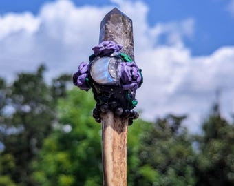 May include: A wand with a clear quartz crystal tip, adorned with purple and green polymer clay flowers and a moonstone cabochon. The wand is made of bone and has a black beaded wrap.