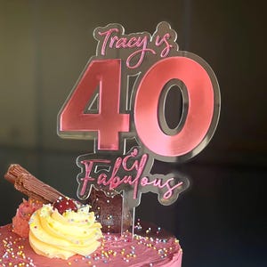 May include: A layered cake with pink, yellow, and brown frosting. The cake is topped with a decorative topper that reads "Tracy is 40 & Fabulous" in pink. The cake is decorated with sprinkles, a swirl of yellow frosting, and a chocolate bar.